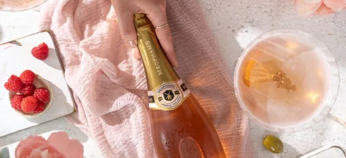 Bottle of Meophams Phoenix sparkling wine on a pink blanket, surrounded by raspberries, pink peonies, candles, and a glass of wine.