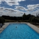 Outdoor swimming pool with bright blue water and clear sky, surrounded by trees and a paved deck with a few people.